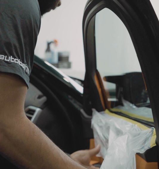 Man installing window tint on a car in a garage. He wears glasses, a cap, and gloves.