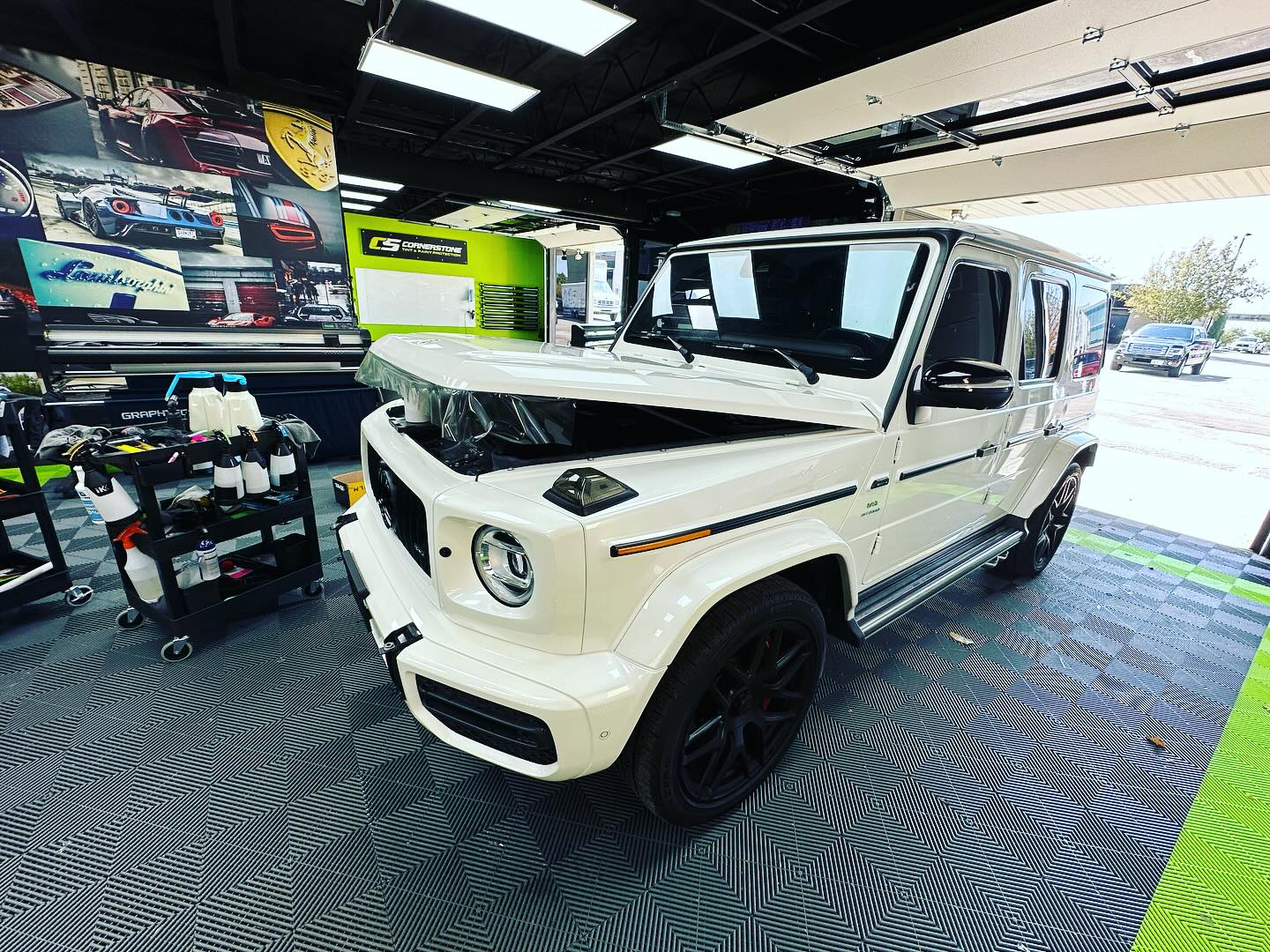White Mercedes G-Wagon in a shop. Hood open, black wheels, on checkered floor, surrounded by detailing supplies.