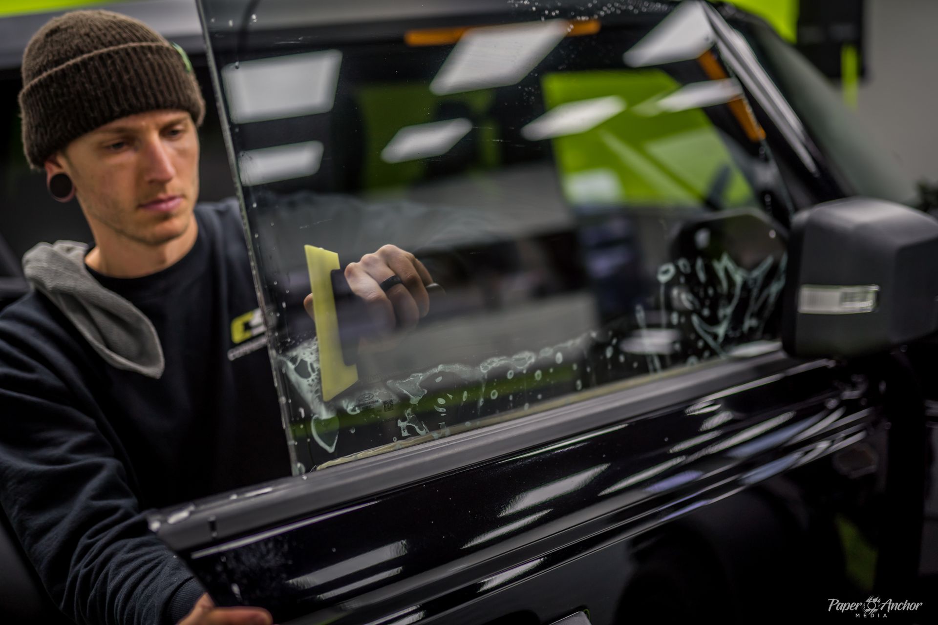 Person applying window tint to a car window. He is wearing a beanie and using a smoothing tool.