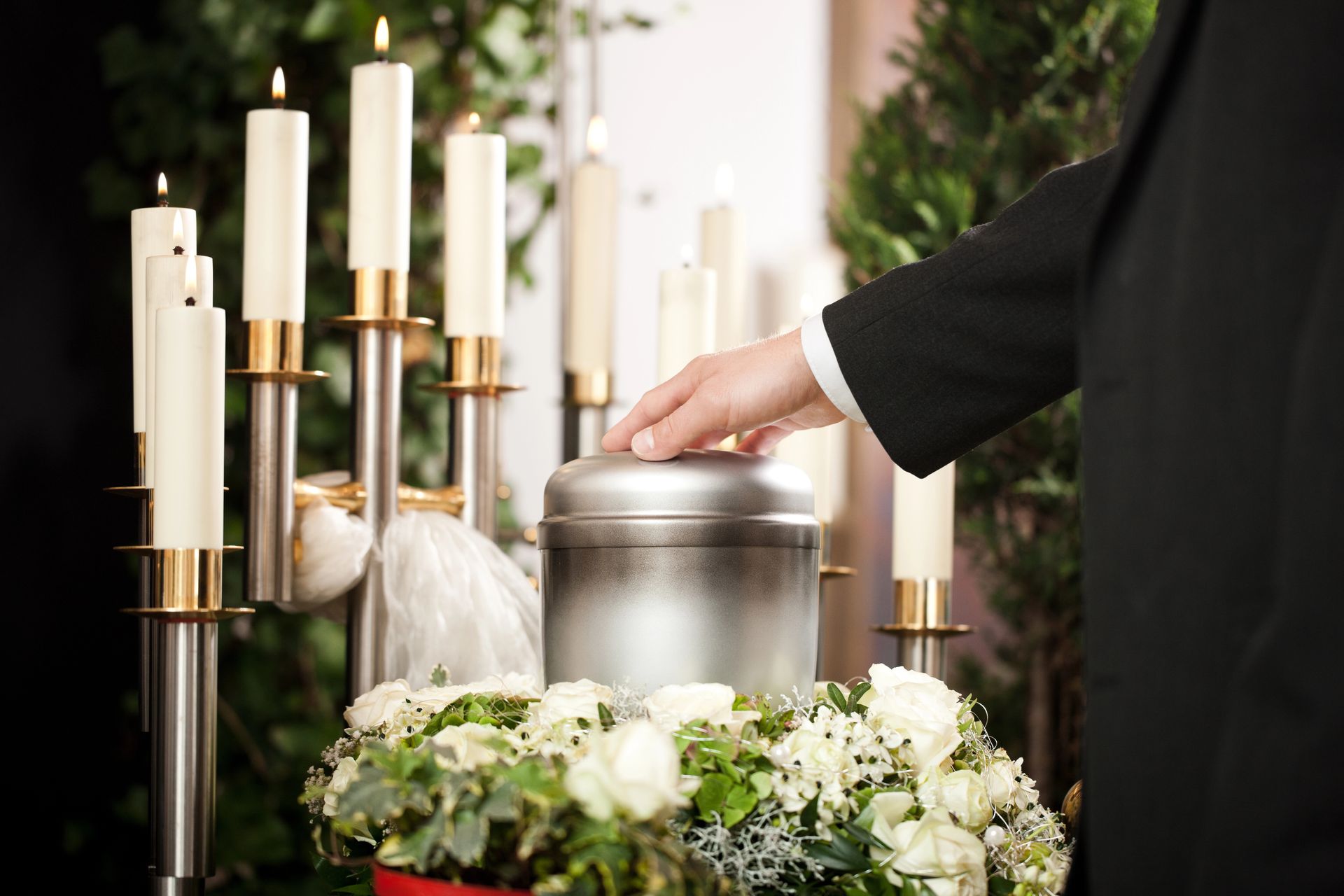 Una mano depone un'urna d'argento accanto a fiori bianchi e candele presso un altare commemorativo.