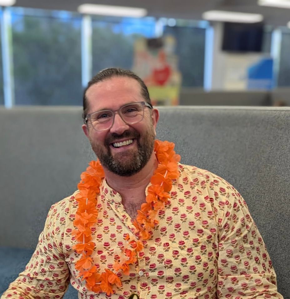 A man wearing glasses and an orange lei is sitting on a couch.
