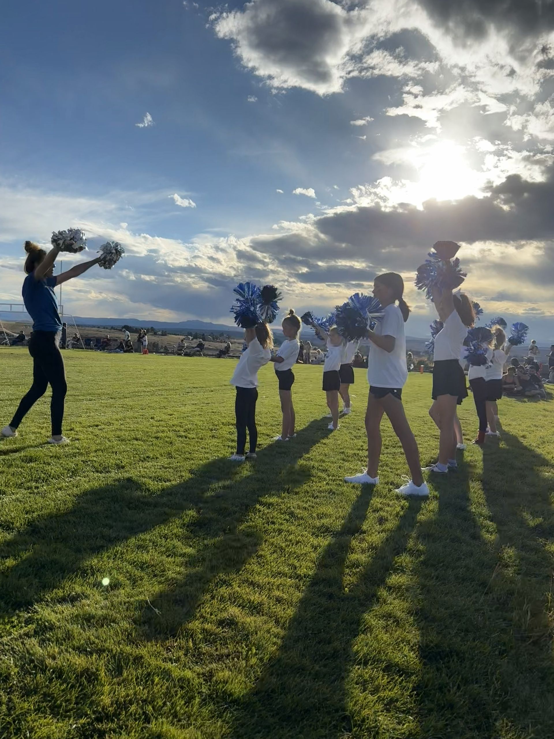 Cheerleaders practice with pom-poms in a grassy field. Blue and white colors. Bright sunlight and cloudy sky in the background.