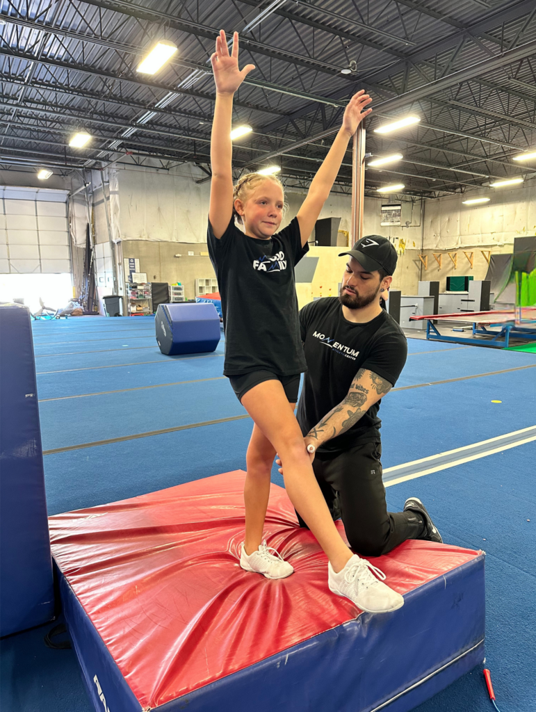 A man is helping a young girl do a trick on a mat in a gym.