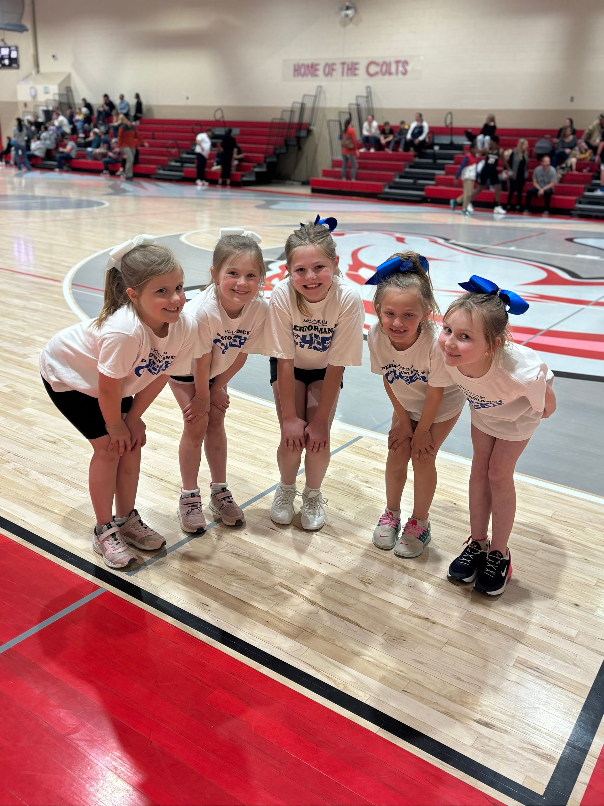 Five young cheerleaders posing on a gym floor, wearing white shirts and blue bows.