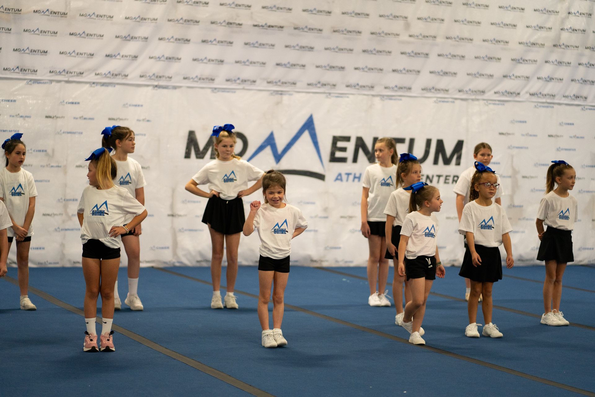 Cheerleaders in white shirts and black skirts, practicing on blue matting in gym; 