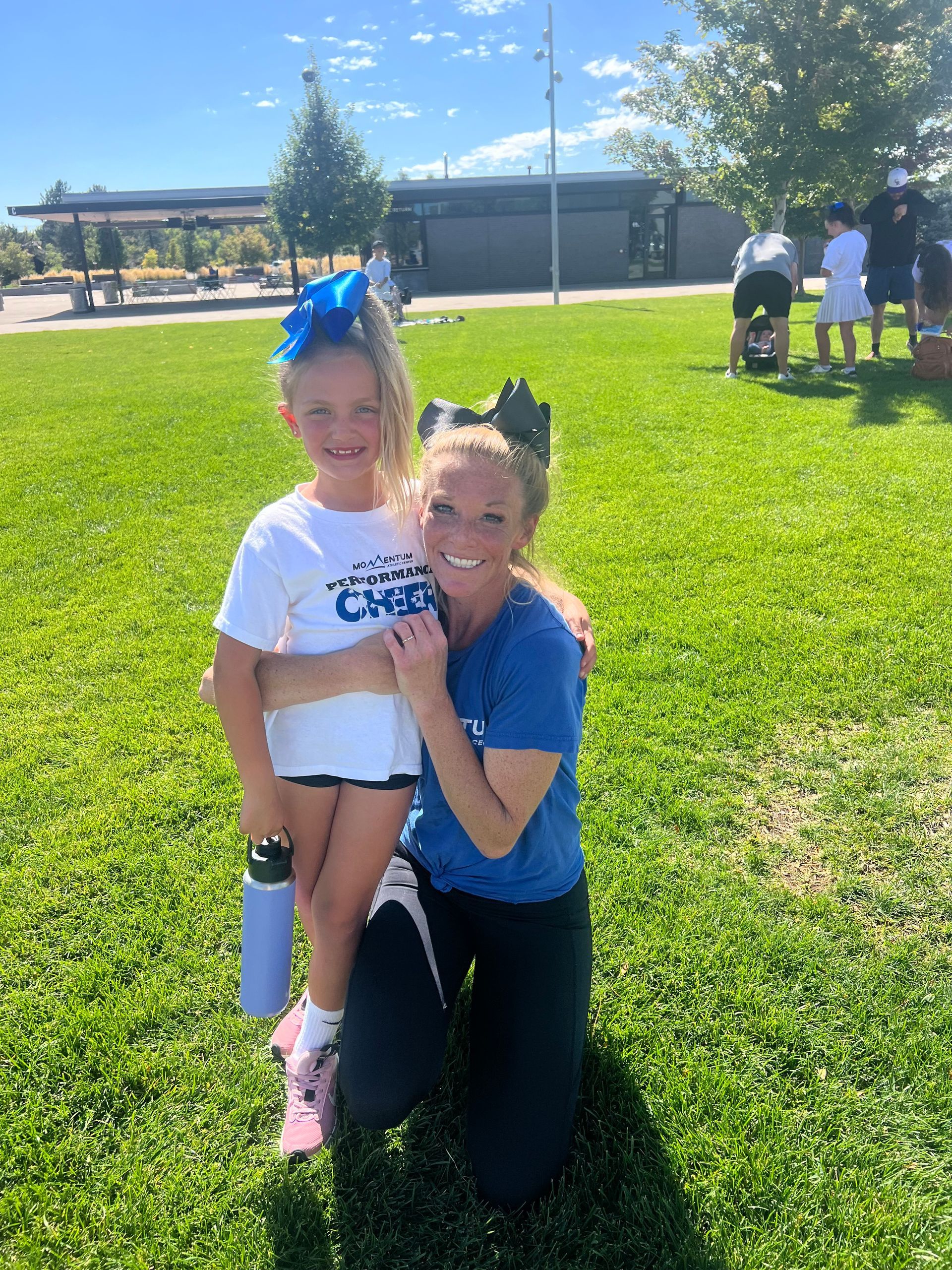 Woman and child in cheerleading attire posing on a grassy field in the sun.
