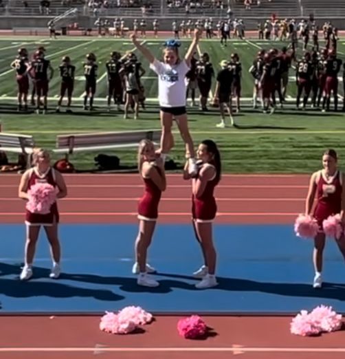 Cheerleaders performing a stunt on a track; one person is held up high. The setting is a football field.