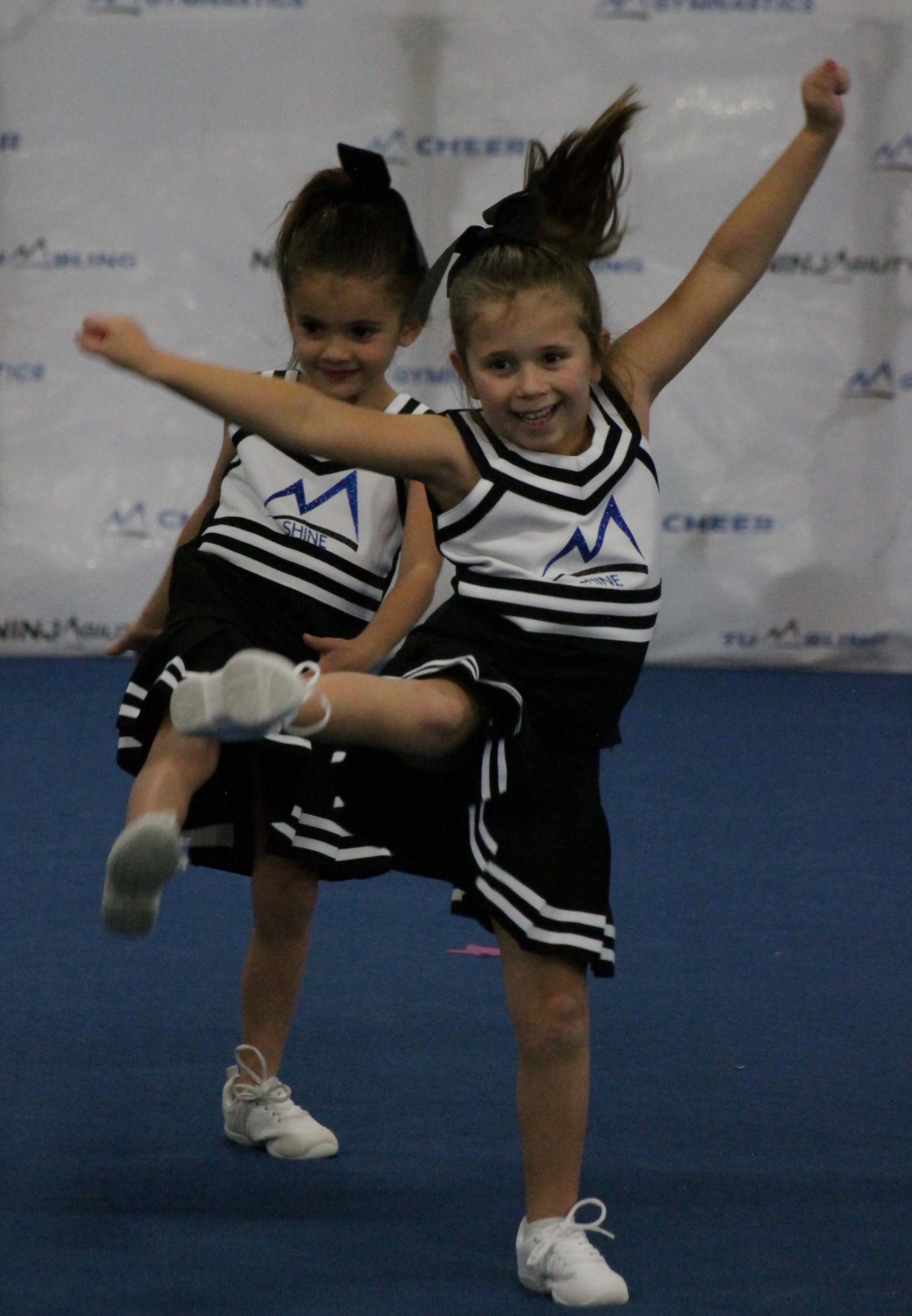 Two cheerleaders in black-and-white uniforms pose mid-routine with arms raised and one leg lifted.