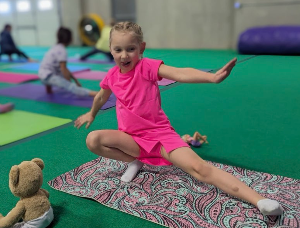 A little girl is sitting on a yoga mat next to a teddy bear.