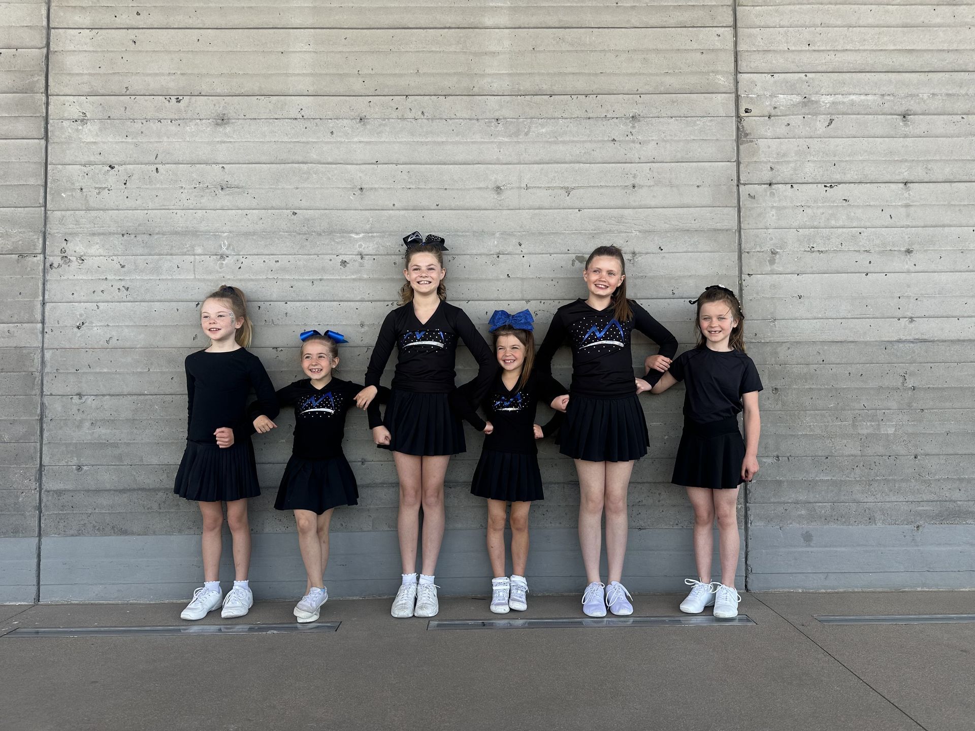 Six cheerleaders in black uniforms stand holding hands in front of a concrete wall.