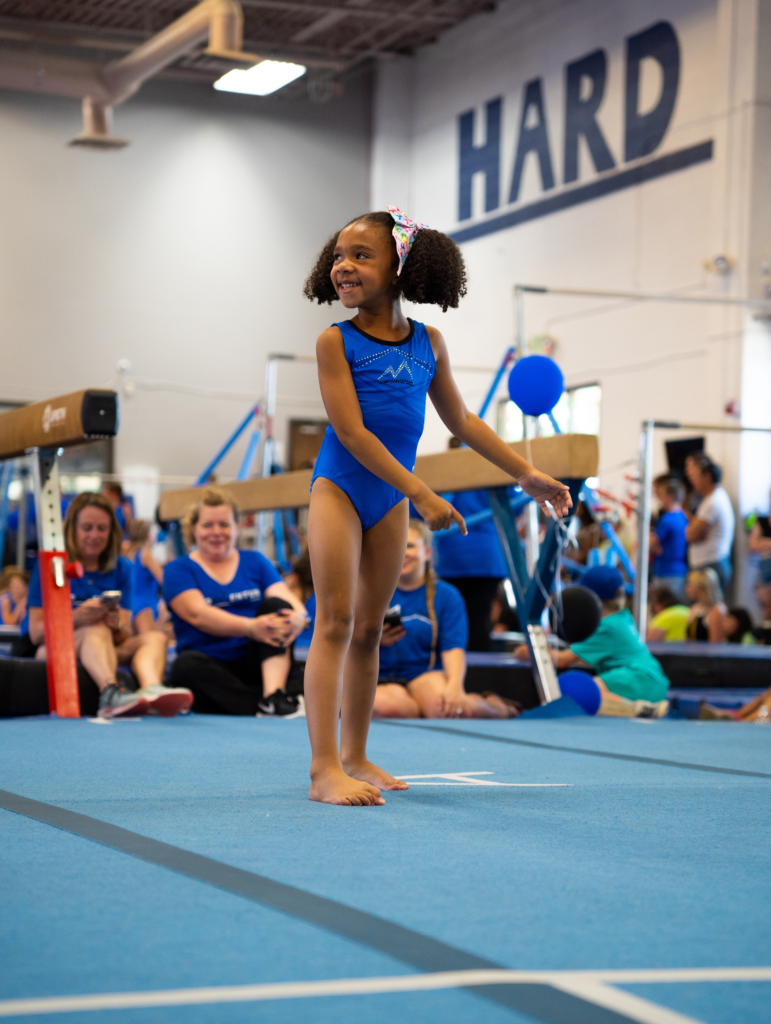 A young girl in a blue leotard is standing on a balance beam in front of a sign that says hard