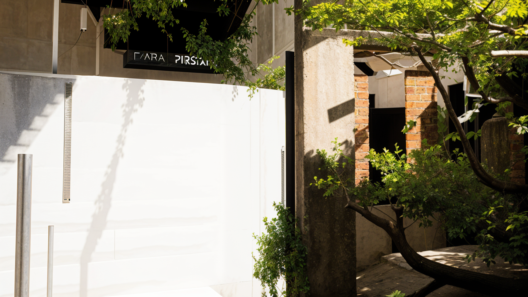 Exterior of a building with white and brown walls, greenery, and partial sunlight.