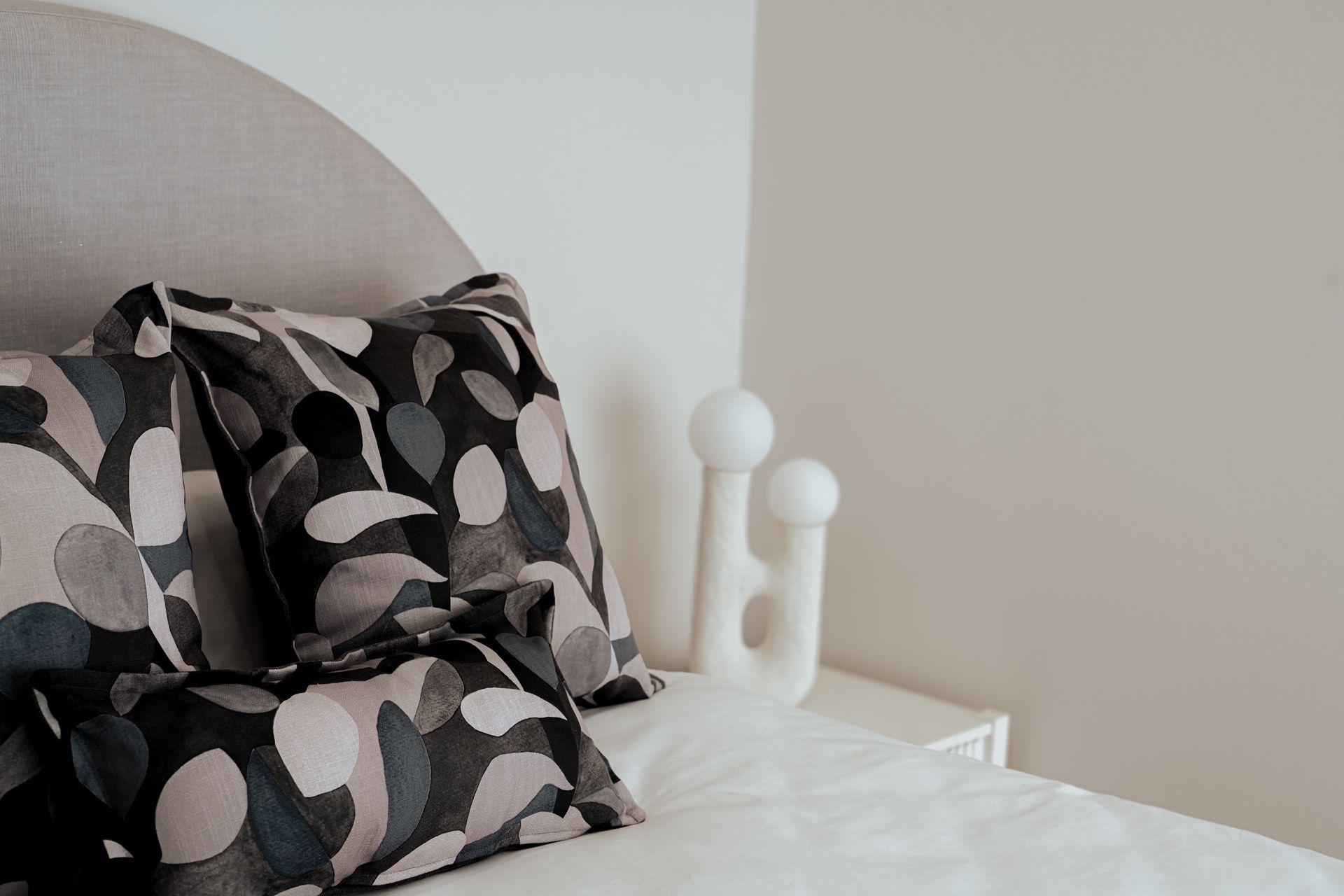 Bed with patterned pillows, headboard, and decorative lamp. White sheets and neutral wall.