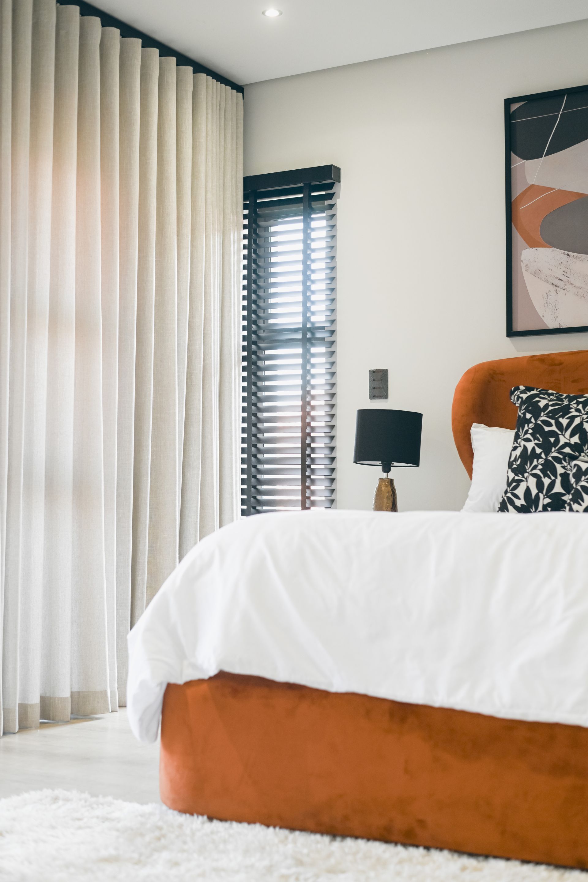 Orange bed with white bedding, black lamp, and window with blinds in a modern bedroom.