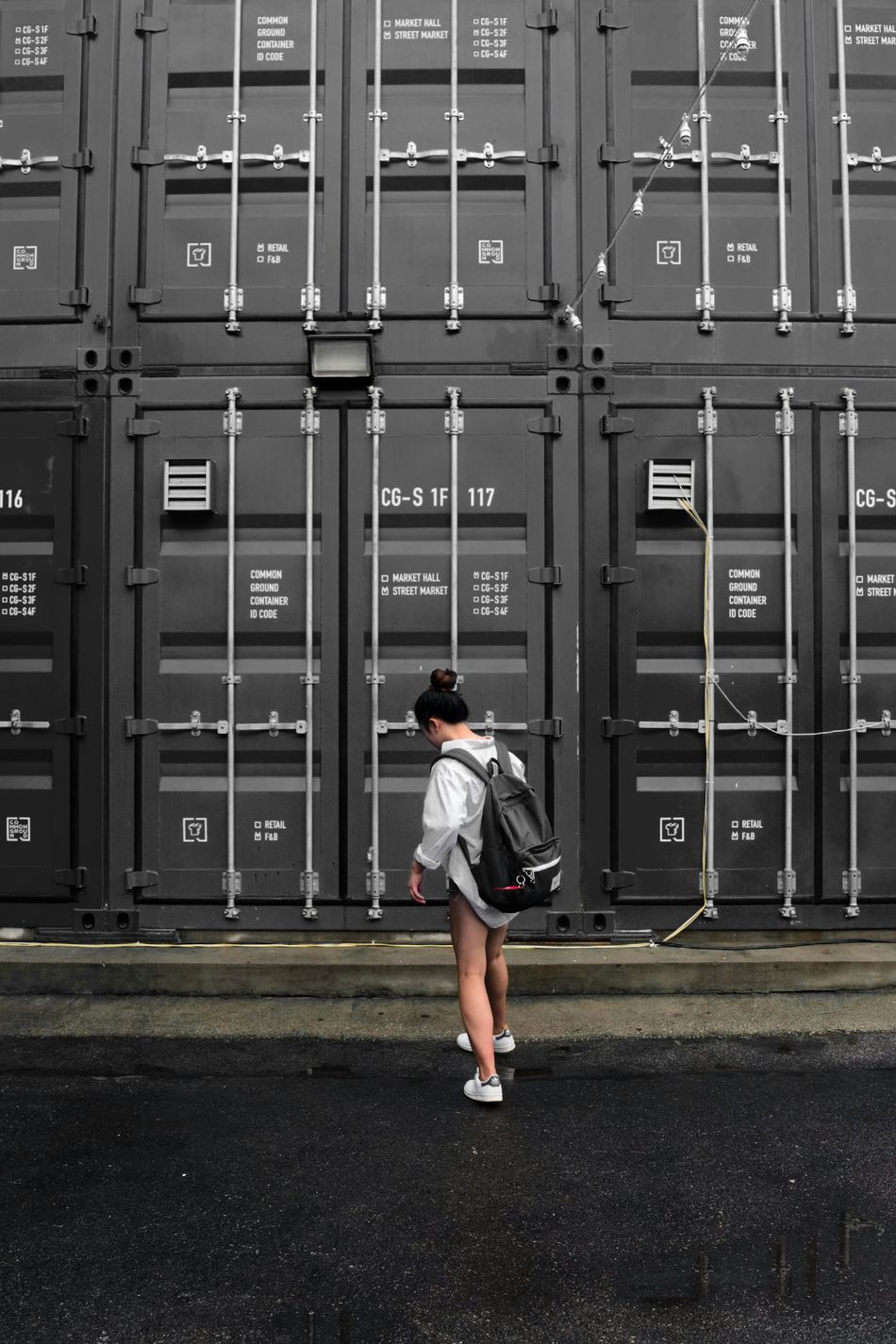 A woman with a backpack is standing in front of a wall of shipping containers