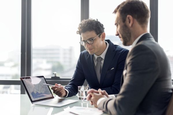 Two men are sitting at a table looking at a laptop computer.