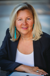 A woman wearing a black jacket and blue earrings smiles for the camera