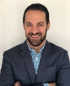 A man in a suit and plaid shirt is smiling with his arms crossed.