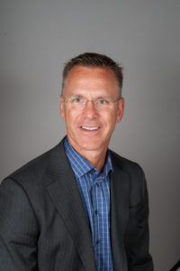 A man in a suit and blue plaid shirt is smiling for the camera.