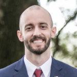 A bald man with a beard is wearing a suit and tie and smiling.