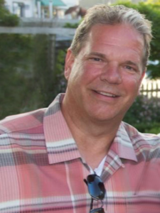 A man wearing a plaid shirt and sunglasses smiles for the camera