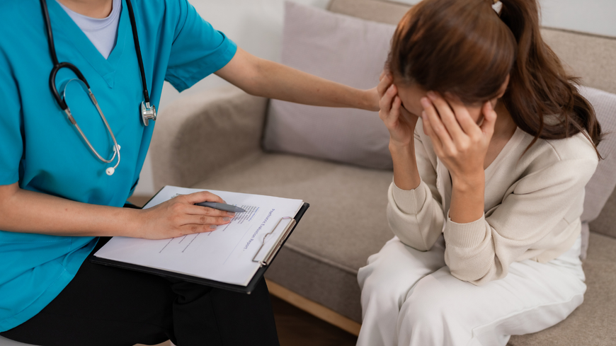 A medical professional comforts a person, holding a clipboard. The person is sitting on a couch, covering their face.