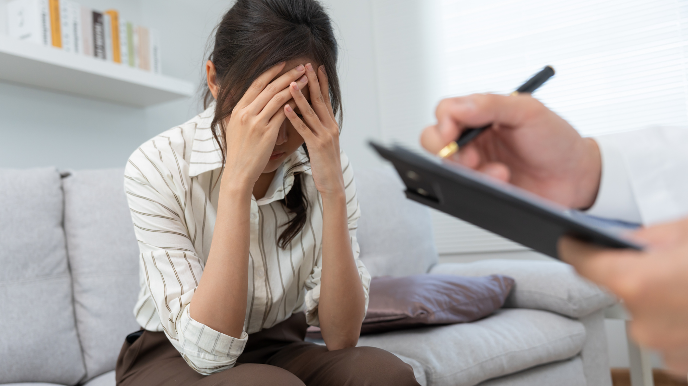 Woman with hands covering her face, sits on a couch while a person writes on a clipboard.