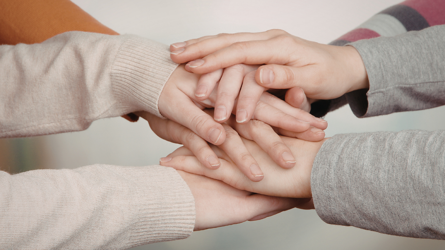 Four people stack their hands on top of each other in a gesture of unity and cooperation.