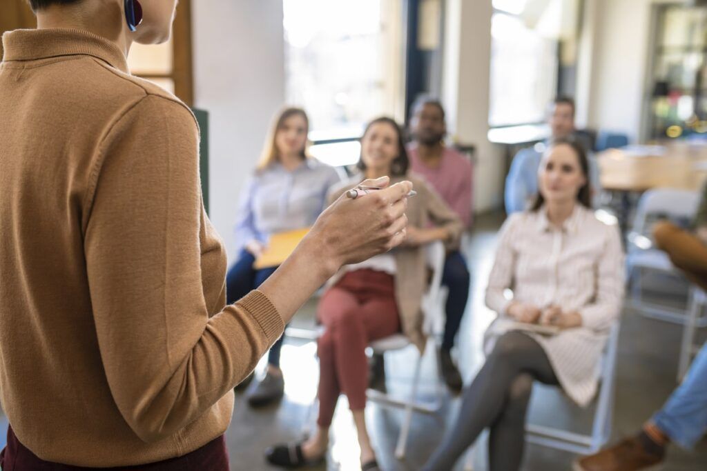 Woman leading a meeting, gesturing with hands. A small group of people are seated, listening attentively.