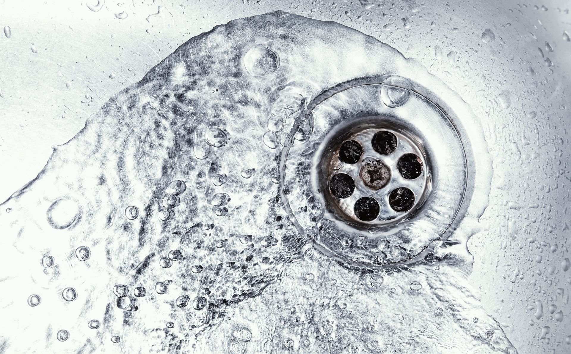 Close-up of water flowing into a metal sink drain with bubbles forming around the opening.