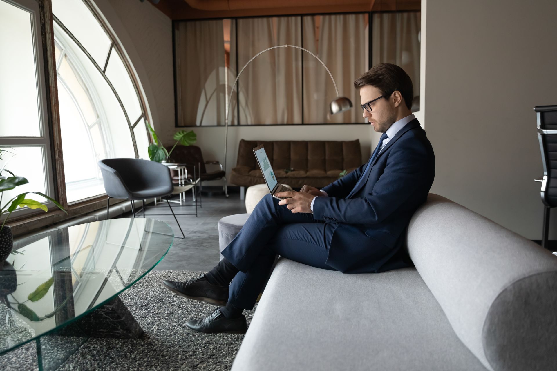 A man in a suit is sitting on a couch using a laptop computer.