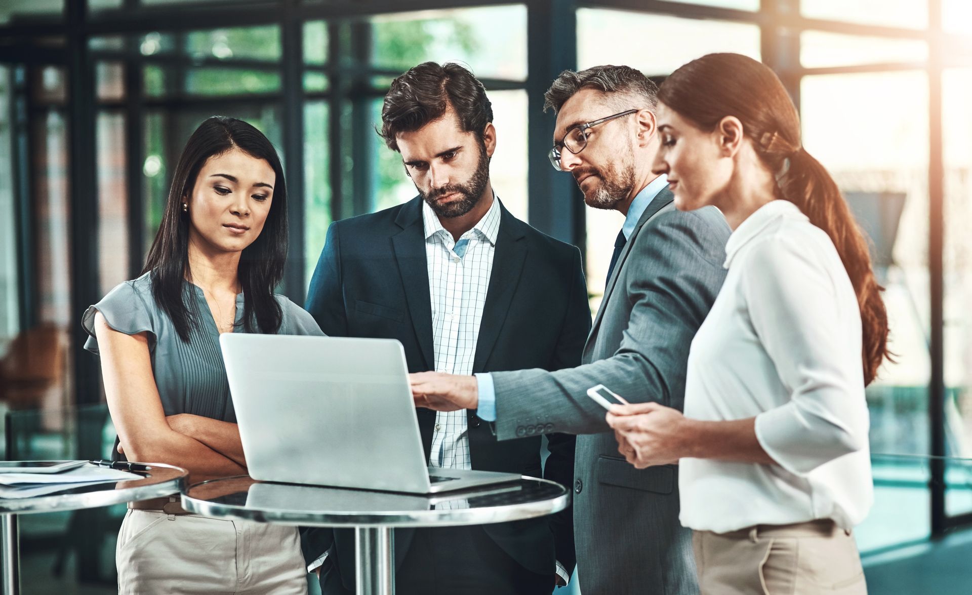 A group of business people are standing around a table looking at a laptop computer.