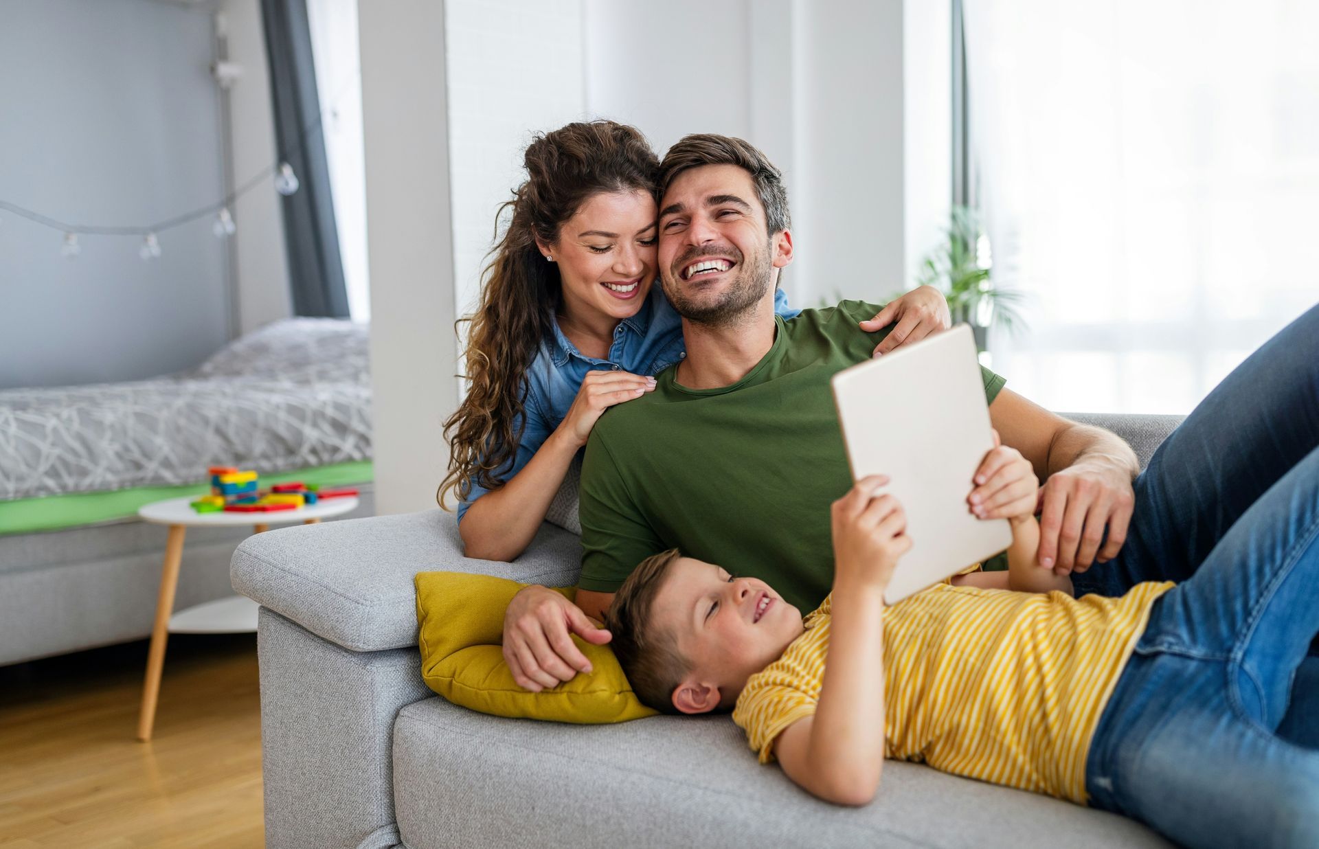 Family relaxing together on a gray sofa, smiling and reading a book in a bright living room