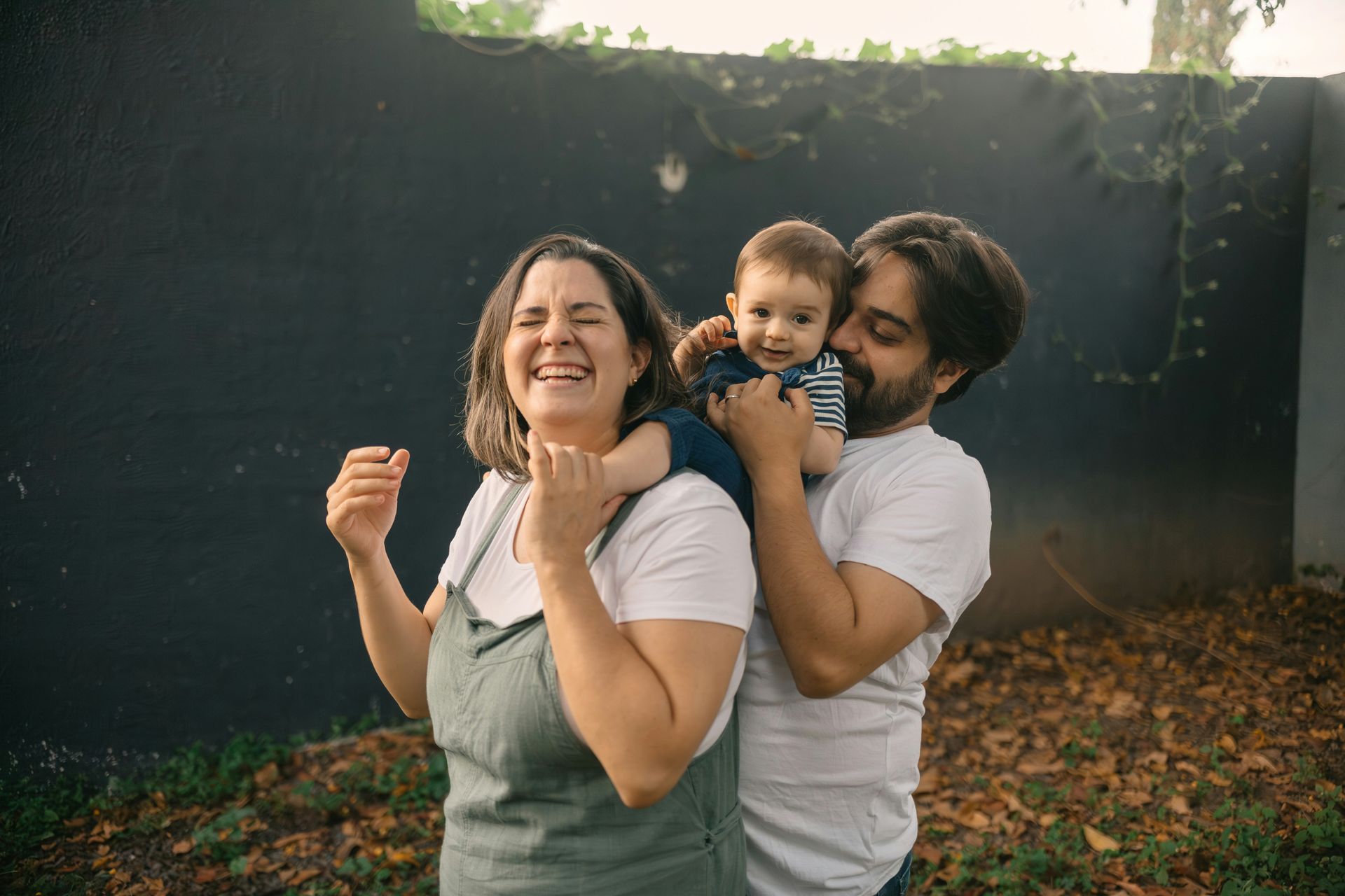 Three people outdoors, smiling and posing in a backyard with a dark fence and autumn leaves.