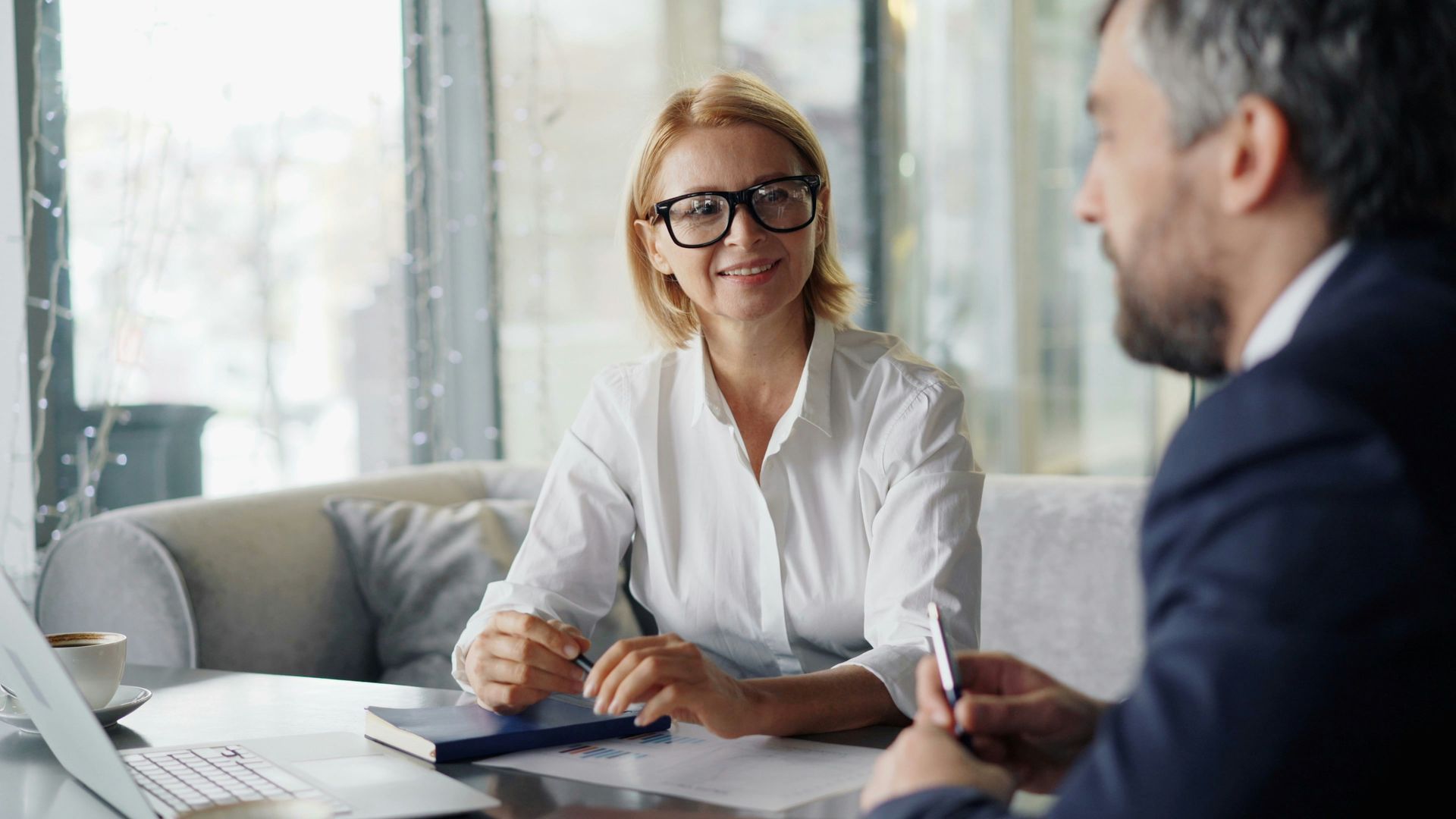 Two people in a bright office meeting, one woman in glasses smiling across the table from a man in a suit