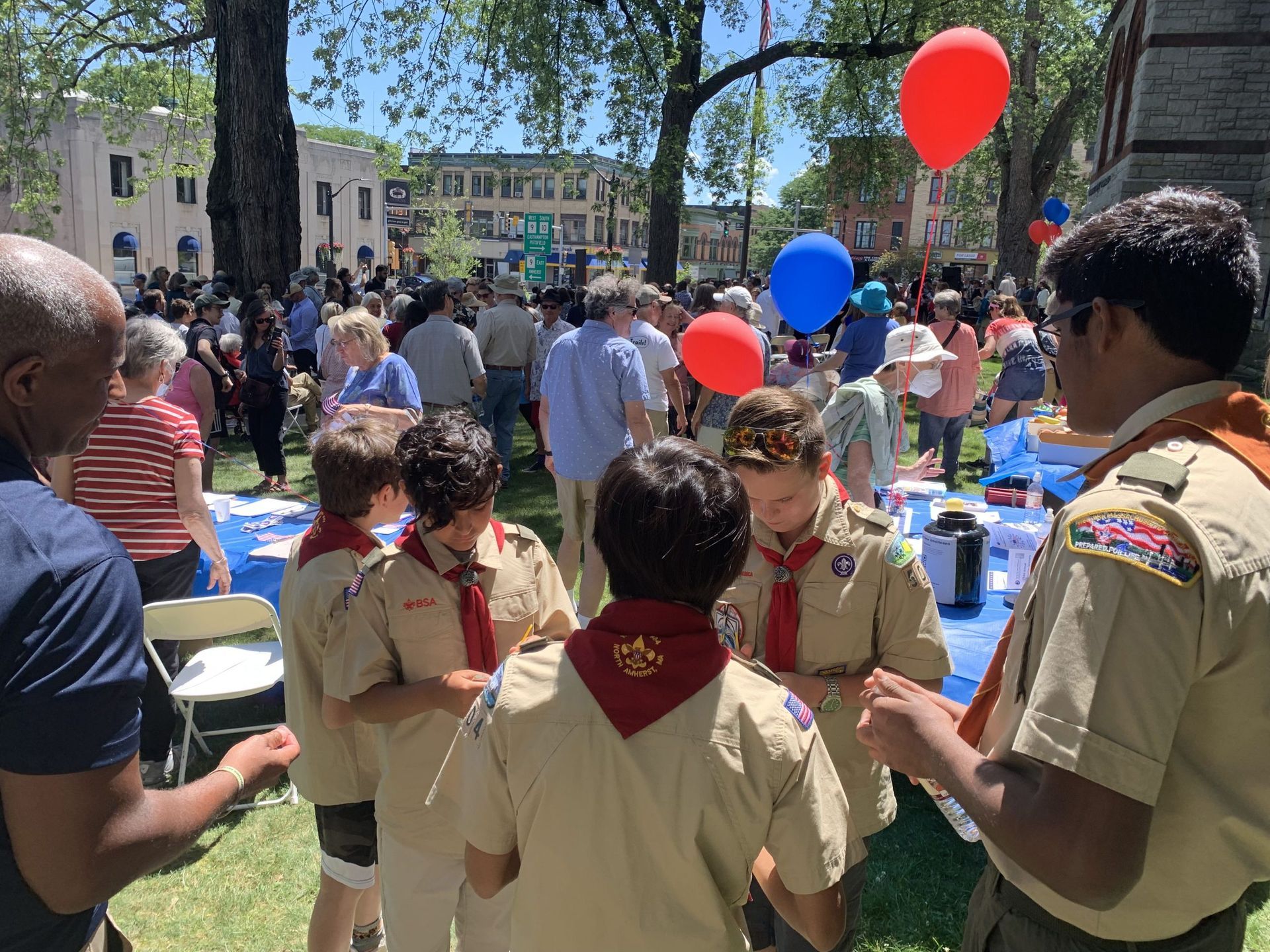 Boy Scouts at a community event. People gather outdoors with red and blue balloons.