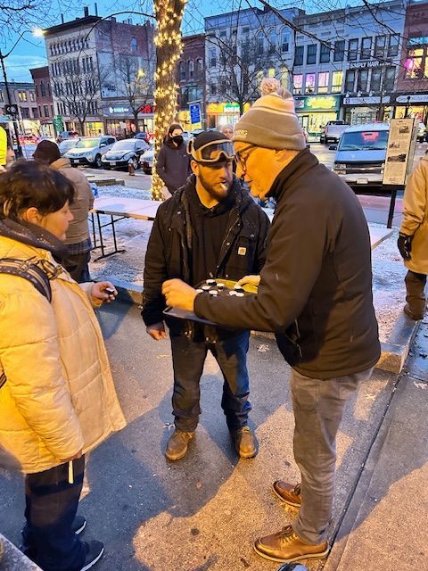 People playing chess on a city street in winter. One person smokes. Buildings and Christmas lights in the background.