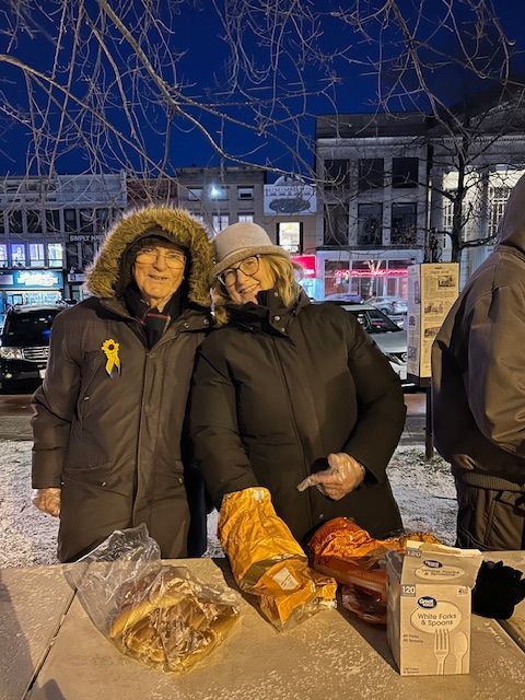 Two people smiling, standing behind a table with bread and food; outdoor, winter setting.