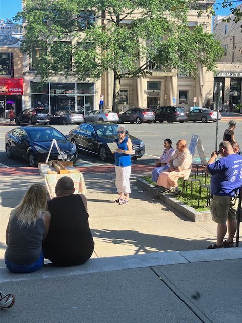 People gathered outdoors. Woman speaking, others seated, painting easel, cars parked on street.