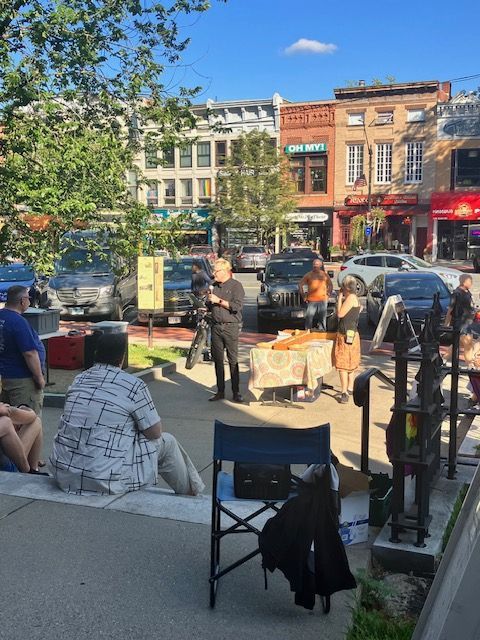 People gathered outdoors in front of storefronts; a man speaks into a microphone.
