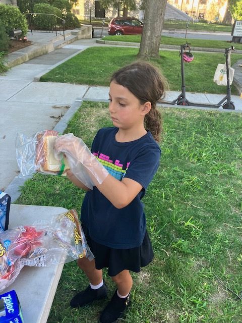 Girl in blue shirt and shorts makes a sandwich outside, near a table of food and grass.