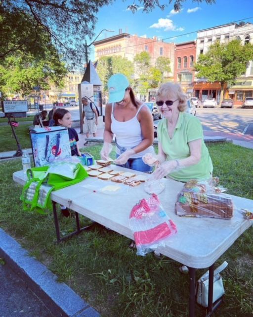 Two women and a child prepare sandwiches at an outdoor table, likely for an event in a park.