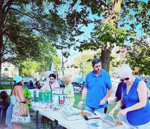 People at an outdoor food stand under trees in a park on a sunny day.