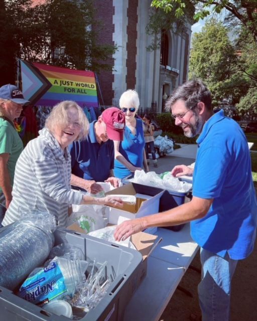 People at an outdoor event distributing food from a table with a rainbow flag in the background.