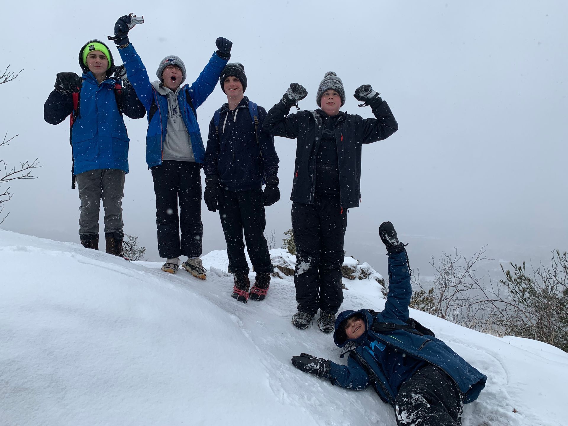 Five people cheering in the snow, one falling. Snowy mountain, gray sky.