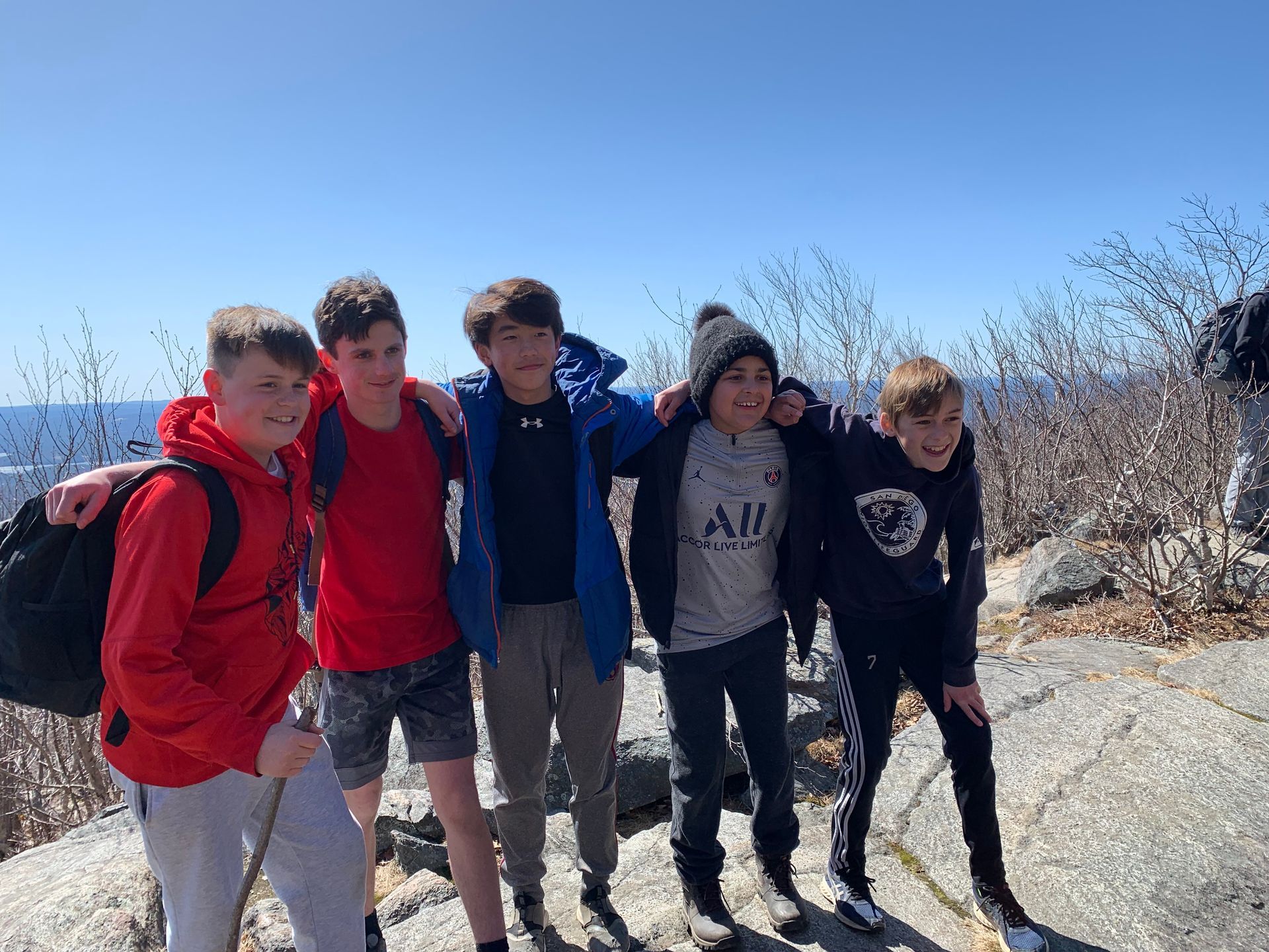 Five boys with arms around each other, smiling on a sunny mountaintop with a coastal view.