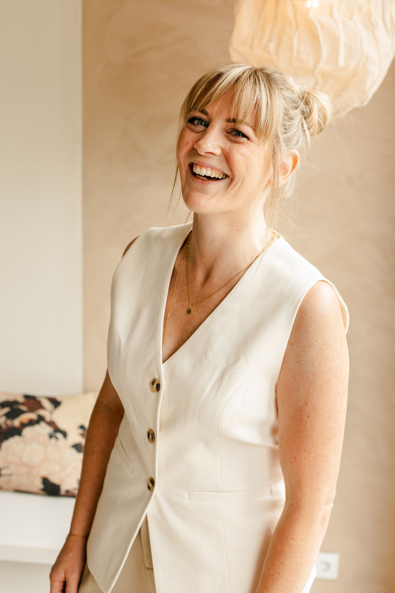 Marielle in crèmekleurig vest glimlacht en staat bij een lichtgekleurde muur en een decoratief kussen.