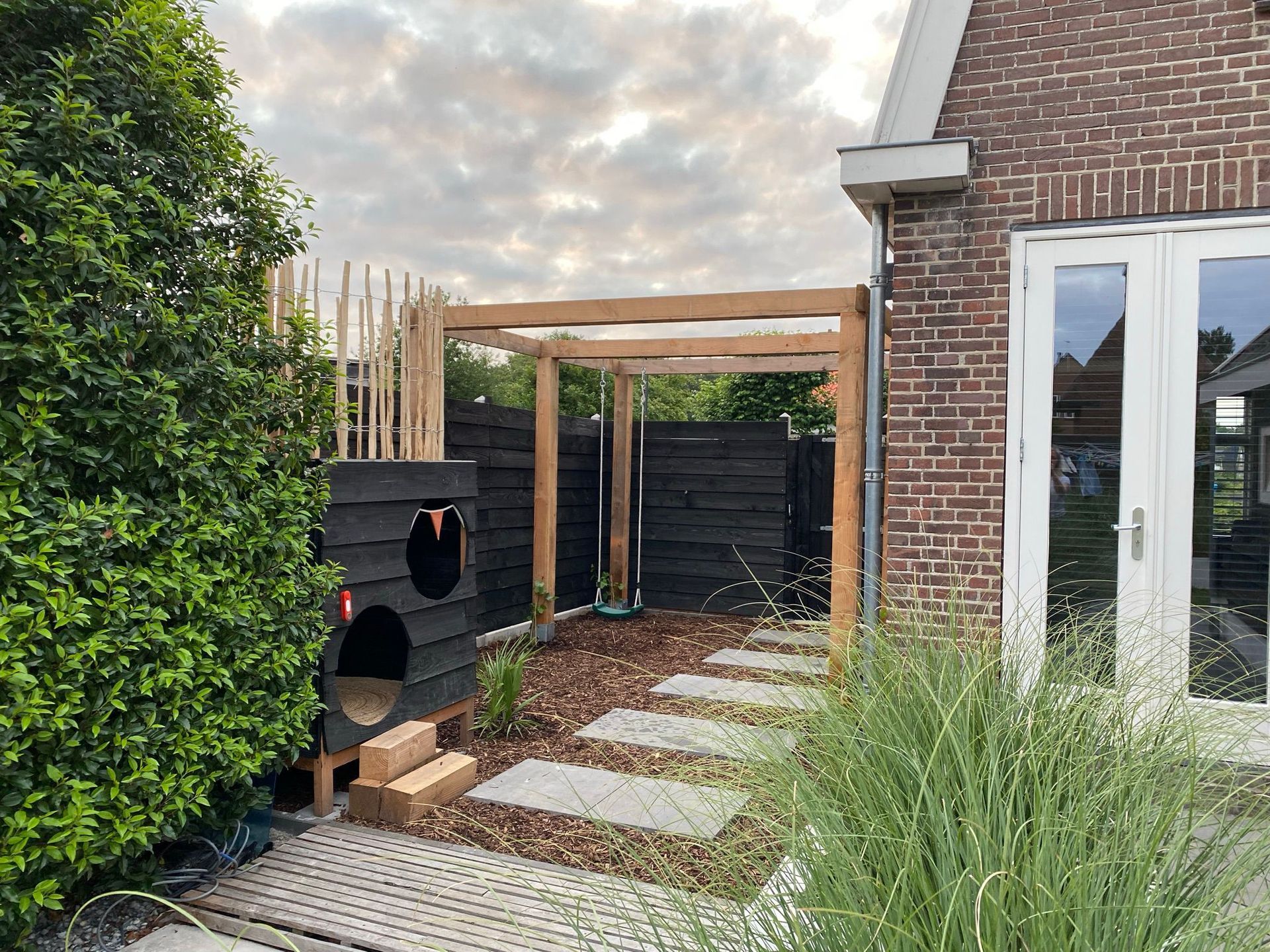 Achtertuin met houten pergola, bakstenen huis, stenen pad en donkere schutting.