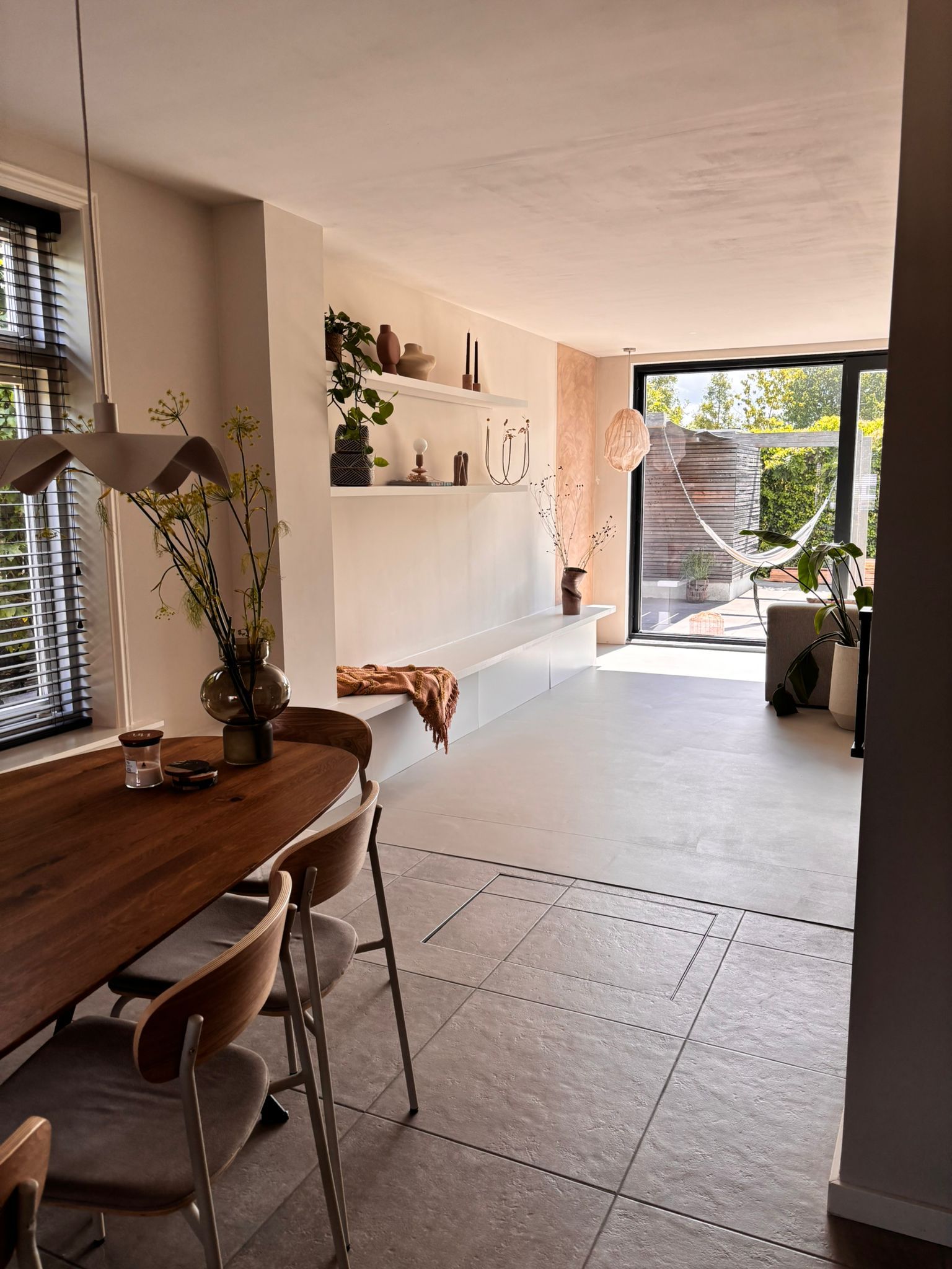 Eetkamer met houten tafel, stoelen, ingebouwde planken en openslaande deuren naar een terras met een hangmat.