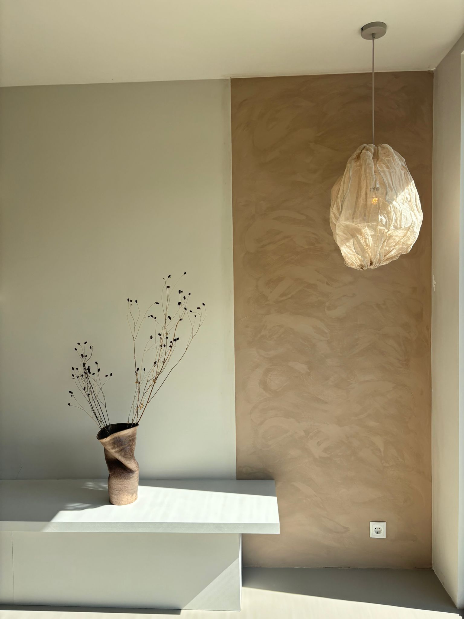 Een beige en lichtbruine muur met een decoratieve vaas en hanglamp in een modern interieur.